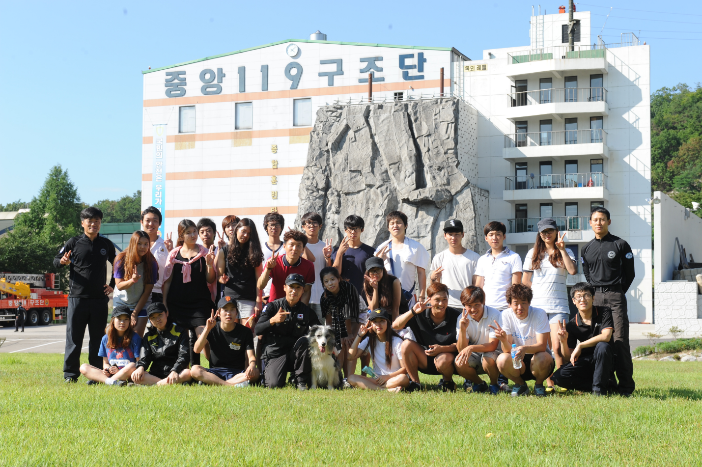 중앙 119 구조단 건물 앞에서 손으로 브이를 그리며 찍은 교수님들과 학생들의 단체 사진. 가운데에 구조견으로 보이는 보더콜리가 있다.
