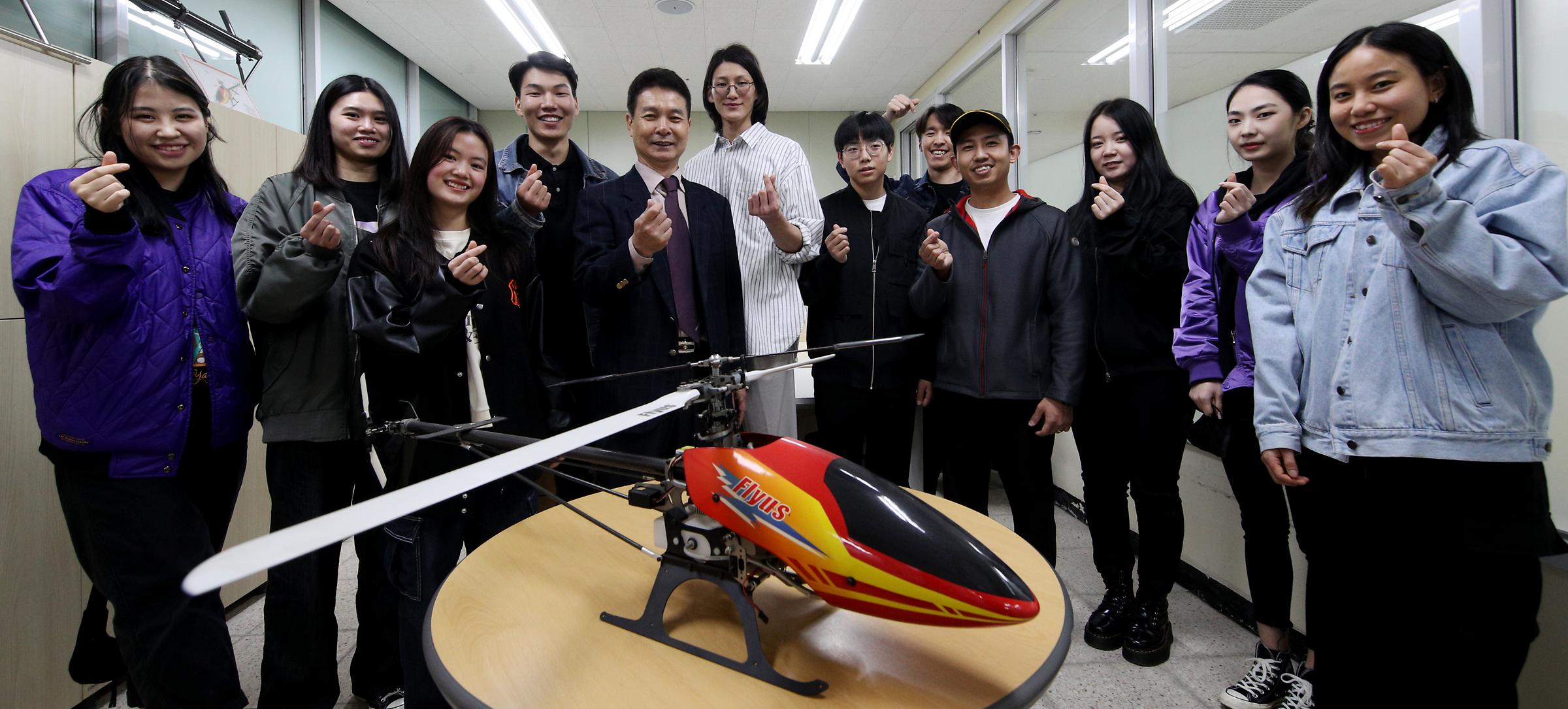 모형 헬리콥터를 앞의 테이블에 올려두고 뒤쪽에 한 줄로 서서 손가락 하트 포즈를 하는 창업경영과 학생들과 교수님
