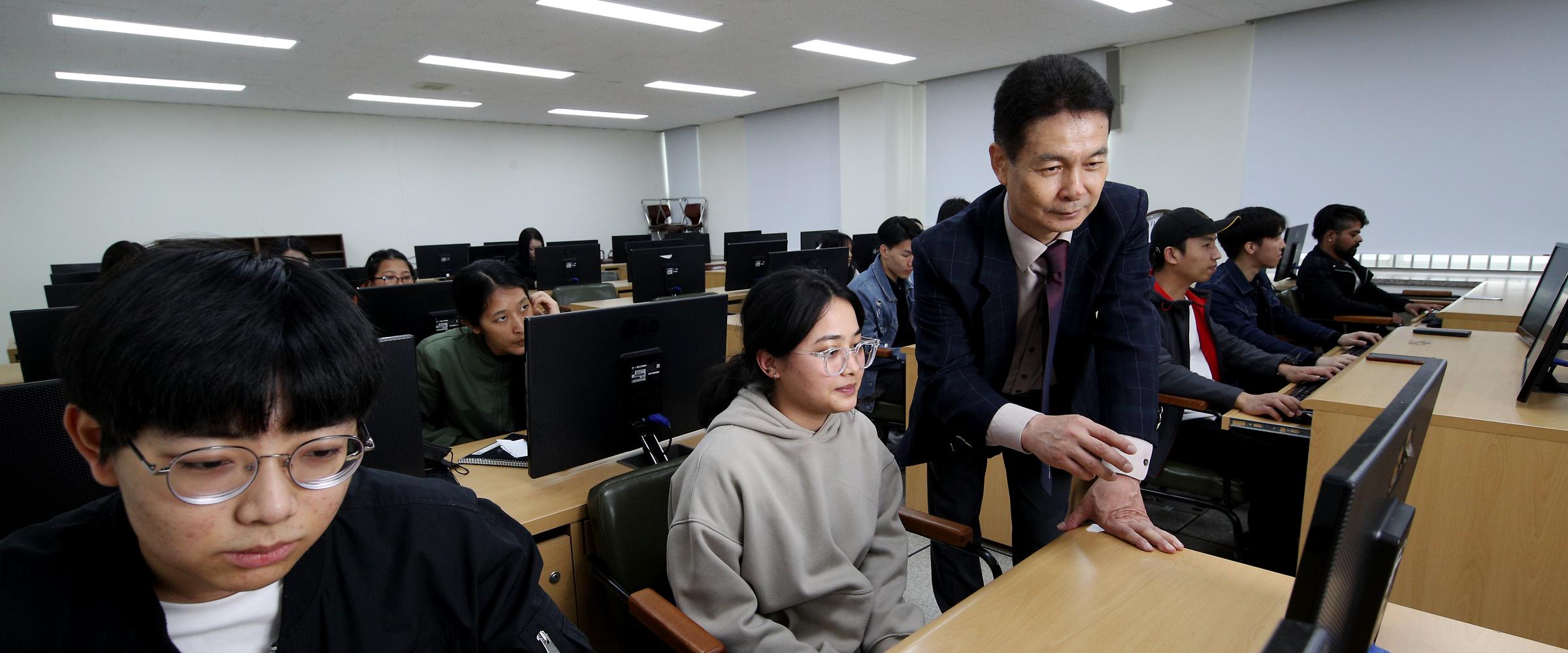컴퓨터 실에서 수업을 듣는 학생들의 사진. 각자 모니터 앞에 앉아 집중하고 있고 한 학생의 컴퓨터 앞으로 교수님이 와 지도 중이다.