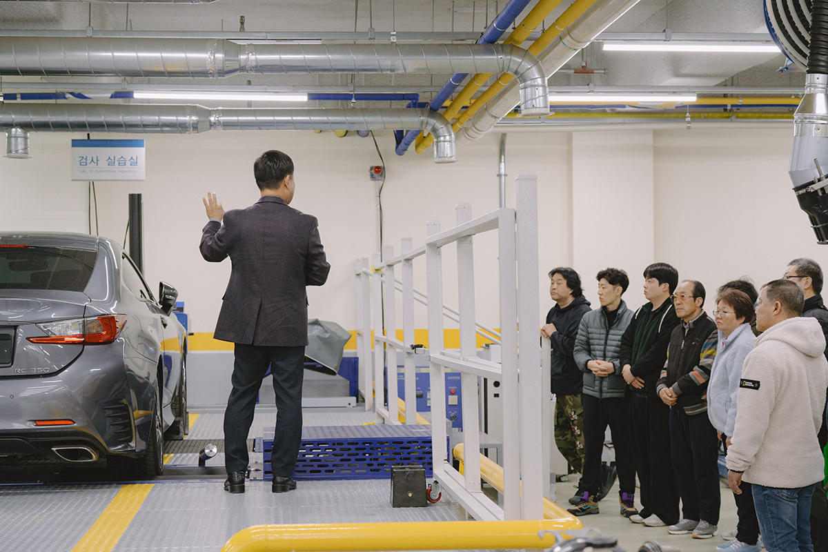 자동차 실습실에서 자동차를 두고, 교수님이 설명하고 학생들은 열심히 듣고 있는 사진