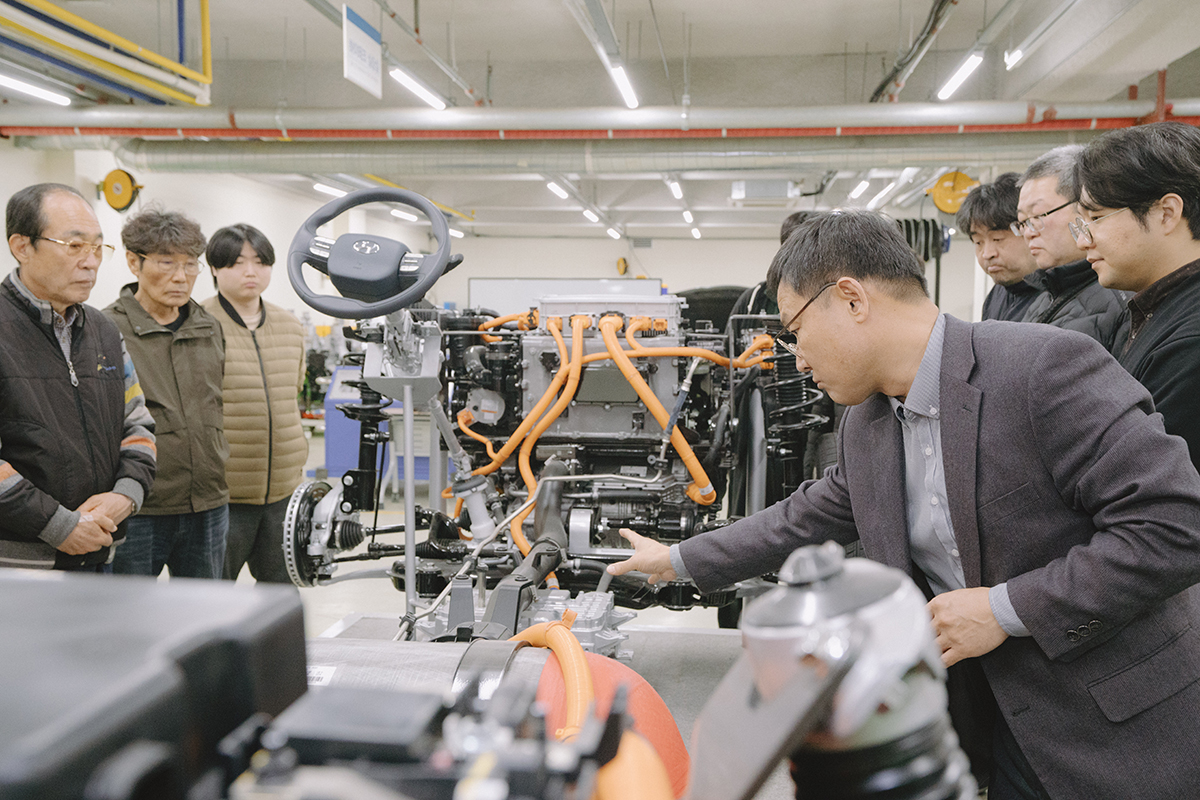 자동차 실습실에서 자동차 후면 쪽 교수님이 설명하고 학생들은 열심히 듣고 있는 사진
