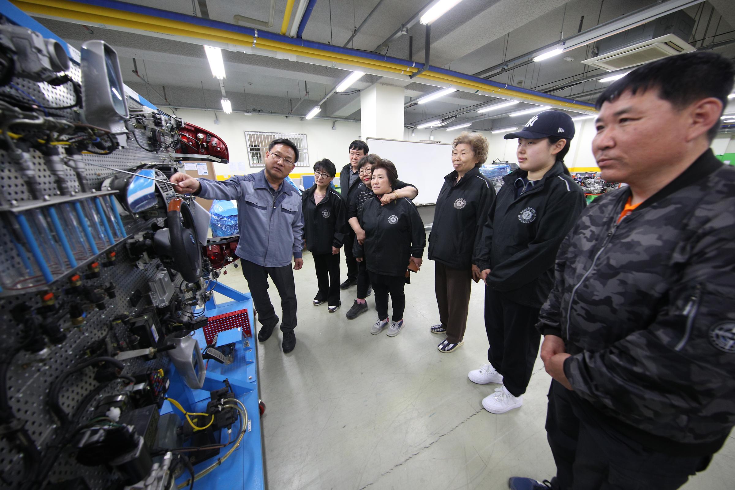 복잡한 자동차 부품을 앞에 두고 설명하는 교수님과 푸품 주변에 둘러 서서 경청하는 학생들